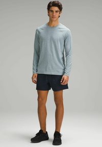 Lichtblauw langemouwen shirt van textuurstof, gecombineerd met een marineblauwe korte broek. Model draagt zwarte sportieve schoenen; achtergrond is neutraal grijs.