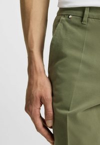 Pantalones verde oliva con tejido texturizado, corte ajustado, bolsillo lateral y un pequeño detalle a rayas cerca de la cintura. Mano apoyada sobre la tela.