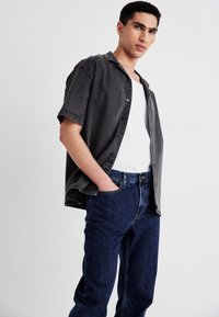 Szürke, rövid ujjú, gombos ing fehér pólóval párosítva, sötétkék farmernadrággal. Az anyag denimszerűnek tűnik, laza fazonnal.