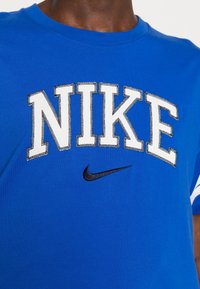 Blå bomullströja med rund hals, med stora vita bokstäver "NIKE" och en svart Nike swoosh-logga nedanför.