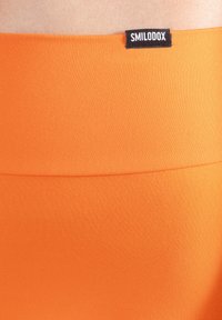 Orange stretchigt tyg med en slät textur, med en svart etikett märkt "SMILODOX" vid midjebandet. Enkel design, inga mönster.