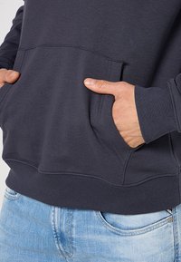 Dunkelblaues Sweatshirt mit Kängurutasche, gerippten Bündchen und Saum, aus weichem Stoff, getragen über hellen Jeans.