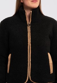 Giacca in pile nera con colletto alto, caratterizzata da accenti beige lungo la zip e le tasche laterali. Esterno texturizzato, fodera liscia.
