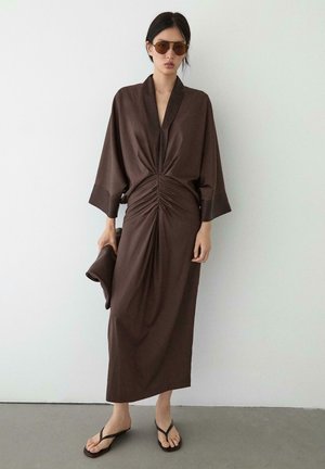 Femme portant une longue robe marron froncée avec de larges manches, tenant une pochette assortie, des lunettes de soleil marron et des tongs noires, debout contre un mur blanc.