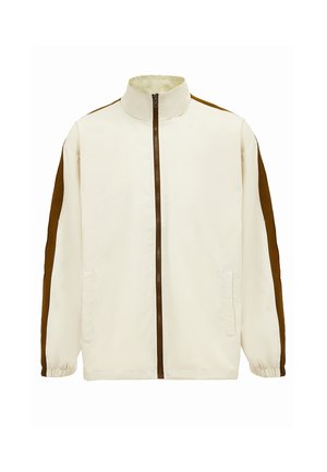 Lys beige jakke med høj krave, brune sidepaneler, fuld lynlås og elastiske manchetter. Har to frontlommer. Glat stofstruktur.