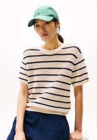 Maglione lavorato a maglia color crema con strisce blu marino, maniche corte e orlo a costine, abbinato a un cappellino da baseball verde, indossato con pantaloni blu marino.