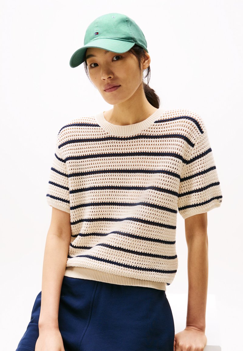 Maglione lavorato a maglia color crema con strisce blu marino, maniche corte e orlo a costine, abbinato a un cappellino da baseball verde, indossato con pantaloni blu marino.