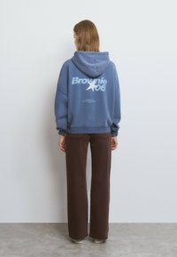 Sudadera azul de gran tamaño con gráfico blanco "Brownie 06" y diseño de estrella, combinada con pantalones anchos marrones y calzado claro.