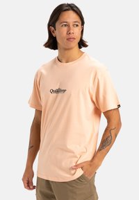 Camiseta de algodón melocotón con mangas cortas, que presenta un logotipo central en negro. Cuello redondo, corte holgado y textura suave.