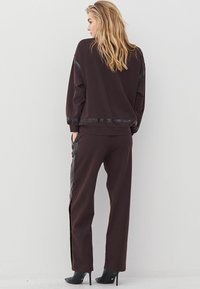 Blondinė moteris stovi priešais baltą sieną, vilkėdama tamsiai rudus laisvo kirpimo kelnes ir derančią ilgą rankovę su juodais akcentais bei aukštakulnius.