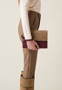 Clutch in pelle marrone a motivo con un accento bordeaux scuro, tenuto contro pantaloni beige testurizzati. Il clutch ha un design a pieghetta con chiusura a scatto.