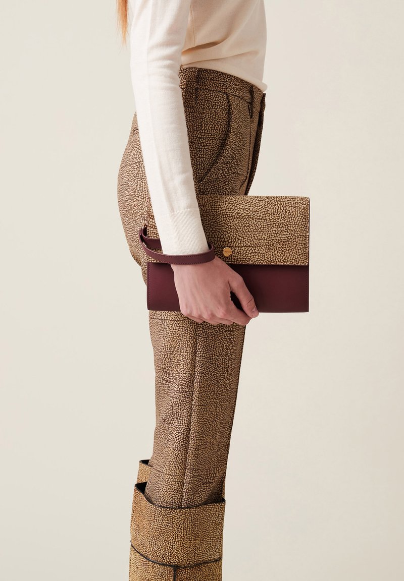 Clutch in pelle marrone a motivo con un accento bordeaux scuro, tenuto contro pantaloni beige testurizzati. Il clutch ha un design a pieghetta con chiusura a scatto.