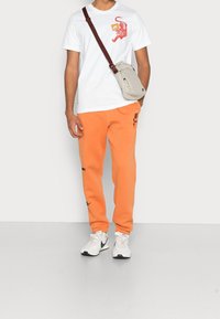 Vit t-shirt med röd tiger grafik, orangea sweatpants med logodetalj, grå crossbody-väska och vita sneakers med svarta accenter.