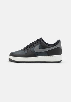 Baskets basses noires et gris foncé avec lacets noirs, logo Nike gris et semelle blanche sur un fond clair uni.