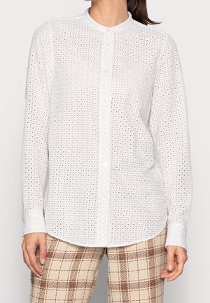 Femme portant un chemisier blanc en broderie anglaise à boutons avec des manches longues et un pantalon beige à carreaux, debout devant un fond uni.