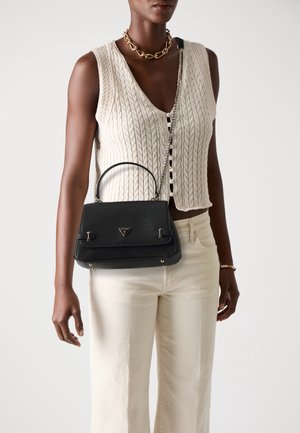 Femme portant un haut beige sans manches en maille torsadée et un pantalon, tenant un sac à main en cuir noir avec une bandoulière chaîne et un collier en or.