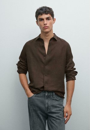 Ung mand med kort krøllet hår iført en mørkebrun skjorte med knapper og optrukne ærmer samt grå jeans, stående mod en lys grå væg.