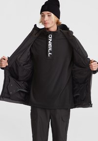 Chaqueta de forro polar negra, desabrochada, con un alto cuello, cremallera frontal y un gran logo de "O'NEILL". Combinada con pantalones cargo negros.