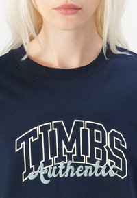 Tamsiai mėlynas džemperis su apvaliu kaklu, su dideliu baltu tekstu "TIMBS" ir mažesniu "Autentiška" šviesiai mėlynos spalvos, pagamintas iš medvilnės, laisvos formos.