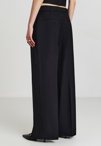 Schwarze, weit geschnittene Hose aus strukturiertem Stoff mit hoher Taille, zwei Gesäßtaschen und klaren Linien. Die sichtbaren Schuhe sind schwarz und spitz.