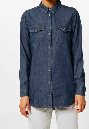 Donkerblauw denim hemd met lange mouwen, knoopsluiting aan de voorkant, twee borstzakken met klep, niet ingestopt gedragen over lichte broeken.