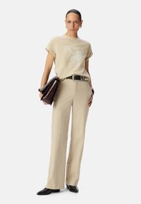 Camiseta gráfica beige con estampado de águila, combinada con pantalones de raya diplomática beige de cintura alta. Cinturón y bolso negros con detalles en cadena. Zapatos negros.