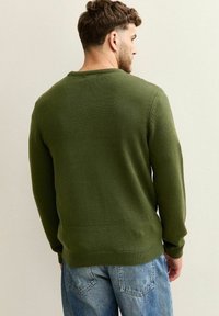 Oliwkowy sweter dzianinowy z okrągłym dekoltem, prążkowanymi mankietami i dołem. Nosi się go z niebieskimi dżinsami, widok z tyłu.