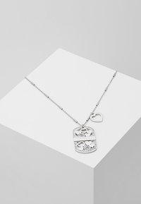 Collier chaîne en argent avec un pendentif rectangulaire comprenant des cœurs découpés, des cristaux et le logo "GUESS", présenté sur un cube blanc.