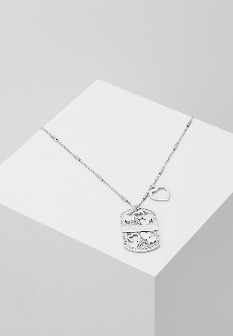 Collier chaîne en argent avec un pendentif rectangulaire comprenant des cœurs découpés, des cristaux et le logo "GUESS", présenté sur un cube blanc.