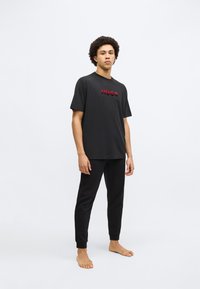 Čierne bavlnené tričko s vyvýšeným červeným logom "HUGO" v páse s čiernymi vzorovanými nohavicami. Zobrazené na bosom jednotlivcovi na jednoduchom pozadí.