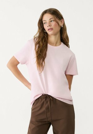 Roze T-shirt met korte mouwen en een ronde hals, gemaakt van zachte stof. Het shirt heeft een ontspannen pasvorm en is gecombineerd met bruine broek met trekkoord.