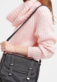 Sac à bandoulière en synthétique noir avec des accents de boucles métalliques doubles, texture lisse et bandoulière réglable, tenu contre un pull en laine rose.