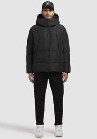 Sort quiltet pufferjakke med høj krave, frontlynlås og to sidelommer. Bæres med sorte cargo-bukser og hvide sneakers.