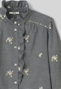 Chemise à carreaux noir et blanc avec un col volants, motifs floraux brodés en blanc et or, et boutons blancs le long de l'avant.