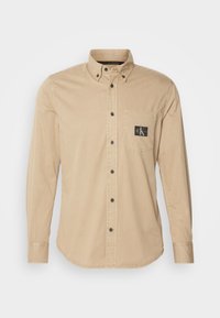 Camicia beige a maniche lunghe con colletto, bottoni neri e taschino sul petto con patch logo. Texture liscia simile al cotone.