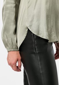 Blouse vert olive clair avec manches longues froncées et ourlet arrondi, associée à un pantalon noir moulant en similicuir.