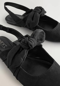 Zapatos de ante negros tipo slingback con un detalle de moño anudado en la punta, con suela plana y correa ajustable para un mejor ajuste.