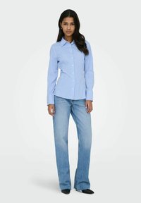 Chemise boutonnée bleu clair avec des rayures verticales blanches, associée à un jean droit bleu clair et des chaussures à bout pointu noires.