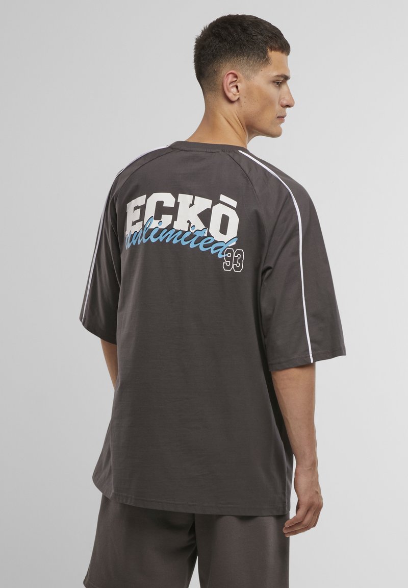 Grå oversized t-shirt med hvid piping, der har et stort grafik i blandet skrifttype på ryggen, som siger "ECKŌ unlimited 93."