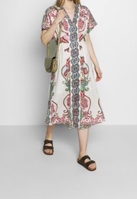 Biała sukienka lniana z kolorowym haftem paisley w odcieniach różu, zieleni i niebieskiego, z dekoltem w kształcie litery V i luźnymi, krótkimi rękawami. Brązowe sandały i zielona torba.