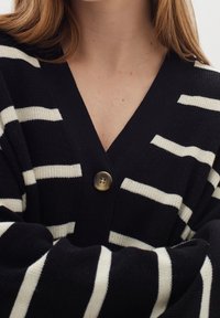 Osoba nosící černobílý pruhovaný kardigan s jedním velkým knoflíkem, s pažemi zkříženými na hrudi.