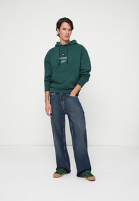 Felpa verde in cotone con tasca frontale e testo bianco, abbinata a jeans blu larghi e sneakers verdi con suola beige.