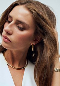 Guldörhängen i form av ringar, ett guldhalsband och ett guldarmband. Släta ytor och glänsande finish mot ljus hud och hår.