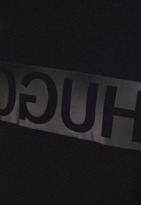 Schwarzes T-Shirt aus Stoff mit einem glänzenden schwarzen Band, auf dem das Wort "HUGO" in fetten, Großbuchstaben über die Brust gedruckt ist.