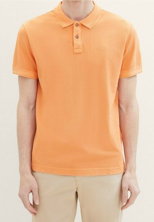 Mand iført en kortærmet orange poloshirt og beige bukser, stående med armene afslappet ned langs siderne mod en neutral baggrund.
