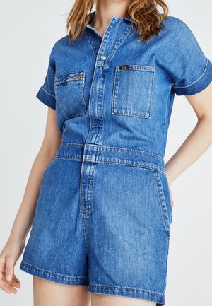 Donna che indossa una tuta corta in denim blu con zip e bottoni sulla parte anteriore, due tasche sul petto e maniche arrotolate, su sfondo bianco.