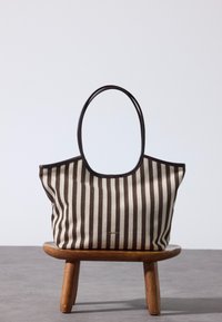 Sac fourre-tout à rayures verticales marron et beige avec poignées foncées, placé sur un petit tabouret en bois devant un fond gris uni.