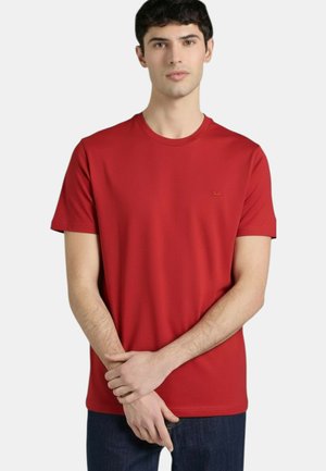 Giovane uomo che indossa una maglietta rossa semplice a maniche corte con scollo rotondo e jeans blu scuro, in piedi con le mani giunte davanti.