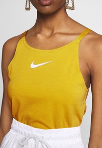 Gul bomullstanktop med tunna axelband och en vit Nike swoosh-logotyp på bröstet. Matchas med vita shorts med elastiskt midjeband.