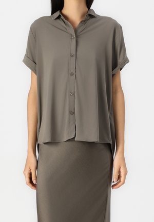 Femme portant une chemise à manches courtes amples à boutons avec les manches retroussées et une jupe cintrée, toutes deux en vert olive pâle.